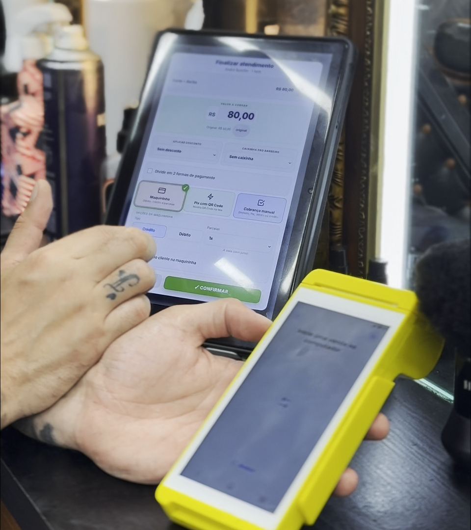 Tablet do Billens na bancada fechando um atendimento de R$ 80,00, com a maquininha amarela na mão do barbeiro.